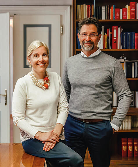 Mag. Šira Fera-Tischler MSc ist am Tisch angelehnt und Mag. Dr. Christof Pollak steht neben dem Tisch, im Hintergrund sieht man Bücher und eine weiße Tür